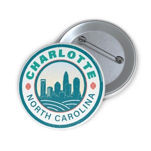 Pu&ograve; includere: Un distintivo rotondo con un design turchese e bianco dello skyline di Charlotte, Carolina del Nord. Il testo "CHARLOTTE NORTH CAROLINA" &egrave; stampato sul bordo. Un secondo distintivo &egrave; visibile sullo sfondo.