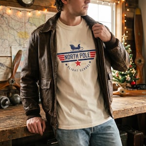 May include: A brown leather aviator jacket and a cream-colored t-shirt with a graphic that reads "North Pole Flight School" in red, white, and blue. The shirt features a sleigh and star design. The person is wearing blue jeans.