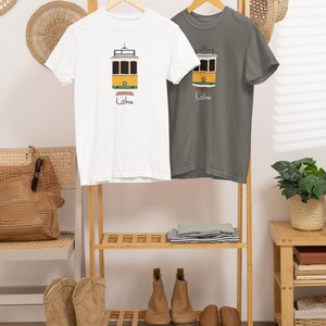 K&ouml;nnte beinhalten: Zwei T-Shirts mit einer Illustration einer Lissabonner Stra&szlig;enbahn. Eines ist wei&szlig;, das andere grau. Die Stra&szlig;enbahn ist gelb und schwarz, darunter das Wort "Lisbon". Die Shirts h&auml;ngen auf einem Holzst&auml;nder mit anderen Kleidungsst&uuml;cken.