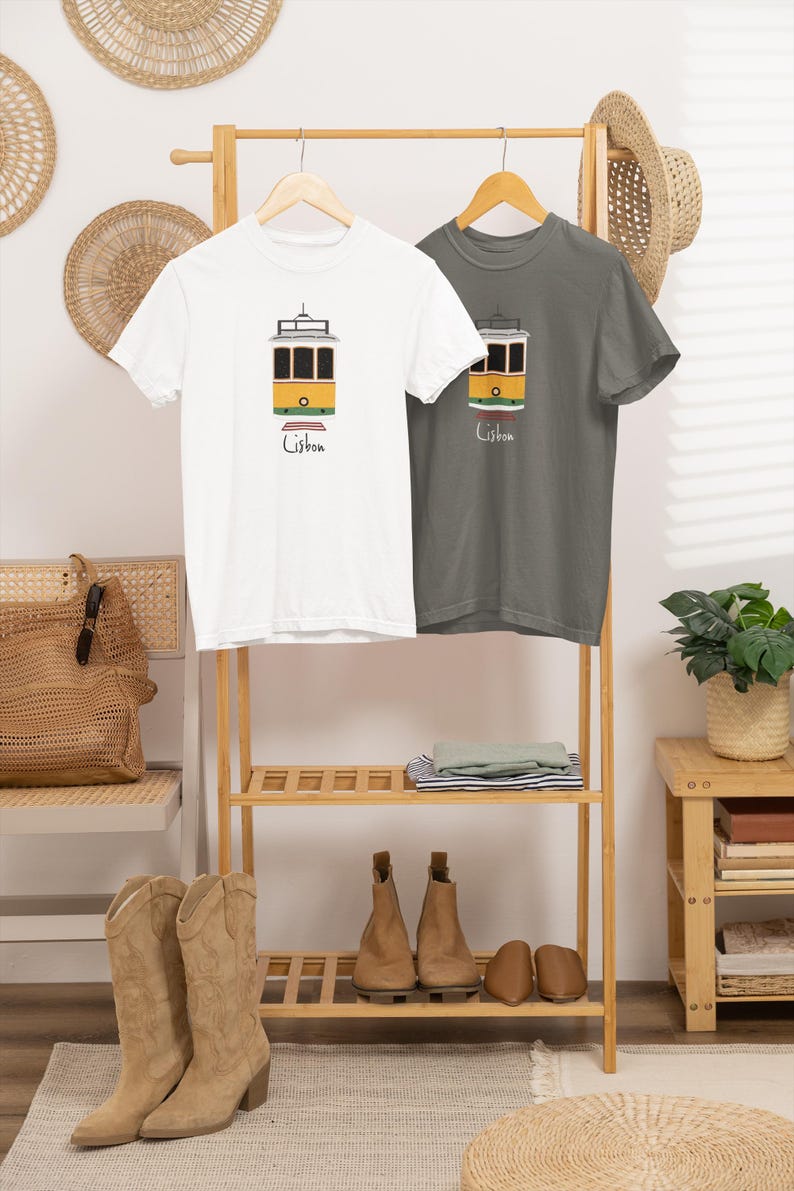 K&ouml;nnte beinhalten: Zwei T-Shirts mit einer Illustration einer Lissabonner Stra&szlig;enbahn. Eines ist wei&szlig;, das andere grau. Die Stra&szlig;enbahn ist gelb und schwarz, darunter das Wort "Lisbon". Die Shirts h&auml;ngen auf einem Holzst&auml;nder mit anderen Kleidungsst&uuml;cken.