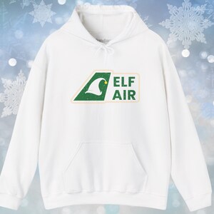 Op de afbeelding: Witte hoodie met een voorzak en trekkoord. De sweatshirt heeft een groen-witte afbeelding met de tekst "ELF AIR" en een elfenmutsontwerp. De achtergrond heeft een sneeuwvlokkenpatroon.