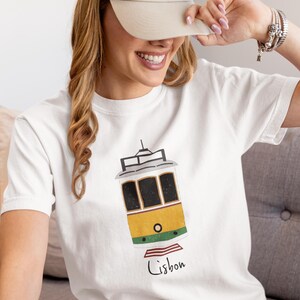 K&ouml;nnte beinhalten: Wei&szlig;es T-Shirt mit einer gelb-gr&uuml;nen Stra&szlig;enbahn-Illustration und dem Wort "Lissabon" in handschriftlicher Schrift. Die Stra&szlig;enbahn hat schwarze Fensterrahmen und ein schwarzes Dach. Die Person auf dem Bild tr&auml;gt eine beige Kappe.