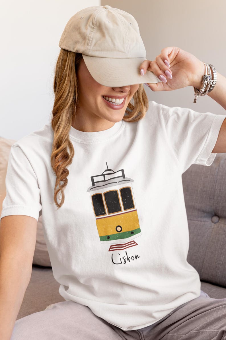 K&ouml;nnte beinhalten: Wei&szlig;es T-Shirt mit einer gelb-gr&uuml;nen Stra&szlig;enbahn-Illustration und dem Wort "Lissabon" in handschriftlicher Schrift. Die Stra&szlig;enbahn hat schwarze Fensterrahmen und ein schwarzes Dach. Die Person auf dem Bild tr&auml;gt eine beige Kappe.