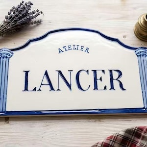May include: A rectangular ceramic sign with a cream-colored background and dark blue lettering that reads ATELIER LANCER. The sign features a decorative arched top and blue column designs on the left and right sides, with a dark blue border.