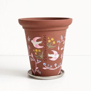 May include: A terracotta-colored ceramic flower pot with a decorative design. The pot features hand-painted pink birds, yellow flowers, and the word "HAPPINESS". It includes a small, round, silver-colored saucer.