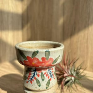 May include: A white ceramic planter with a floral design featuring red and green flowers. The planter has a wide, round top and a narrow base.