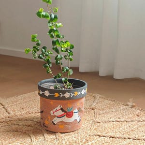 May include: A terracotta-colored ceramic plant pot with a carousel horse design. The pot has a dark gray rim and a decorative band with floral and leaf patterns. The pot contains a green leafy plant. The pot sits on a woven jute mat.