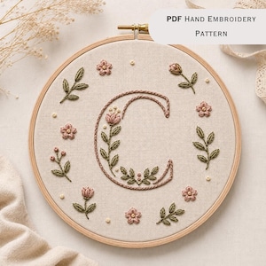 May include: A circular wooden embroidery hoop containing beige fabric with a hand-stitched letter C in pink and green thread. The letter is decorated with floral motifs, leaves, and small buds. Text at the top reads PDF HAND EMBROIDERY PATTERN.