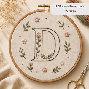 May include: A circular wooden embroidery hoop containing a cream-colored fabric with a hand-stitched letter D. The letter is outlined in dark thread and decorated with embroidered pink flowers and green leaves. Text reads PDF Hand Embroidery Pattern.