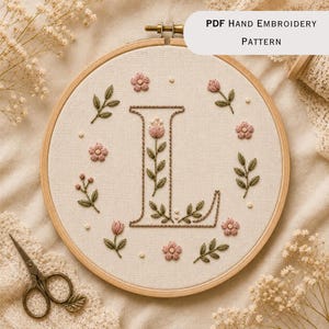 May include: A circular wooden embroidery hoop containing off-white fabric with a hand-stitched letter L in brown thread, surrounded by small pink floral motifs and green leaves. The text PDF Hand Embroidery Pattern is visible at the top.