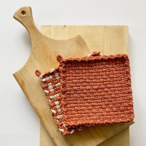 May include: Two woven pot holders with a white and orange checkered pattern. The pot holders are on top of a wooden chopping board.
