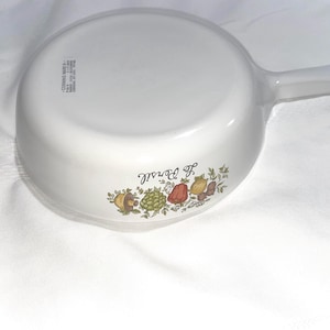 May include: White ceramic saucepan with a black handle and a floral design featuring mushrooms, peppers, and herbs. The text "The  of  " is printed on the bottom of the pan.