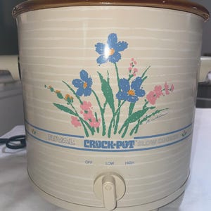 May include: Cream-colored Rival Crock-Pot slow cooker with a floral design featuring blue and pink flowers. The appliance has a brown rim and a clear lid. The control knob has settings for off, low, and high.