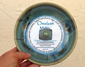 May include: Ceramic omelette maker with a teal and brown glaze. The circular dish has a label that reads "Omelette Maker" and includes instructions. A unique kitchen tool for making omelets.