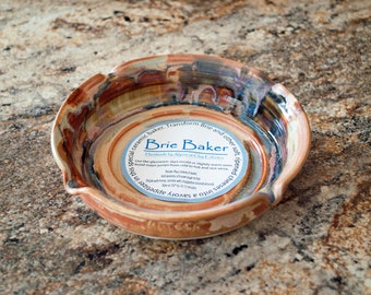 May include: A ceramic Brie Baker with a brown, orange, and blue glaze. The baker has a round shape with a slightly raised rim. The baker has a label that reads "Brie Baker Handmade by Aspen at Clay Creations".