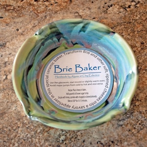 May include: A green and blue ceramic brie baker with the text "Brie Baker" and instructions for baking brie cheese. Handmade by Alyssa at Clay Eclectics.