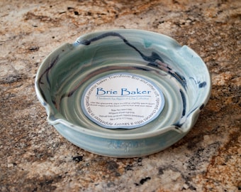 May include: A light blue ceramic brie baker with a white label that reads "Brie Baker Handmade by Algen at Clay Eberhart Use like glassware, start accent on slightly warm oven Avoid major jumps from cold to hot and vice versa Bake at 325 degrees for 10-15 minutes"