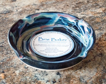 May include: A blue and white ceramic brie baker with a scalloped edge. The baker has a white label with the text "Brie Baker Handmade by Alena & Joy Edwards Use like glassware, start cold or at slightly warm oven. Avoid major jumps from cold to hot and vice versa."