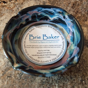 May include: A dark blue ceramic baking dish with a white rim and a colorful glaze. The dish is labeled "Brie Baker" and has instructions for baking brie cheese.