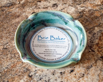 May include: A handmade ceramic Brie baker with a teal and blue glaze. The baker has a scalloped edge and includes a recipe for baked brie. The text on the label reads "Brie Baker" and "Handmade by Alyana at Clay Collective."