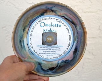 May include: A ceramic bowl with a blue, brown, and white glaze. The bowl has a round opening in the center with a white label that reads "Omelette Maker" and "Handmade by Alyans at Clay Eclectics".