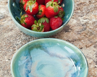 May include: A teal ceramic bowl filled with fresh red strawberries with green leaves. A matching empty bowl sits below. The bowls have a glossy finish and are on a speckled countertop. The bowls are likely for serving fruit.