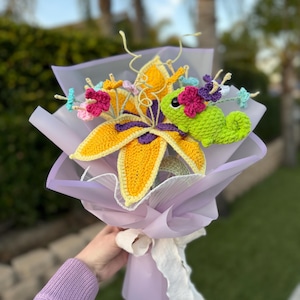 May include: A crocheted bouquet of flowers with a yellow lily and a green chameleon. The bouquet is wrapped in lavender tissue paper and tied with a white ribbon.