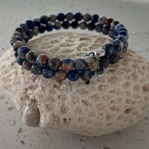 May include: A double-strand bracelet made of blue, brown, and orange gemstone beads. The bracelet is secured with a silver clasp. The bracelet is resting on a light-colored, textured stone.