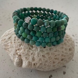 May include: A stack of turquoise beaded bracelets with a silver accent piece. The bracelets are made of small, round beads in varying shades of green and blue. The bracelets are resting on a textured, off-white stone.