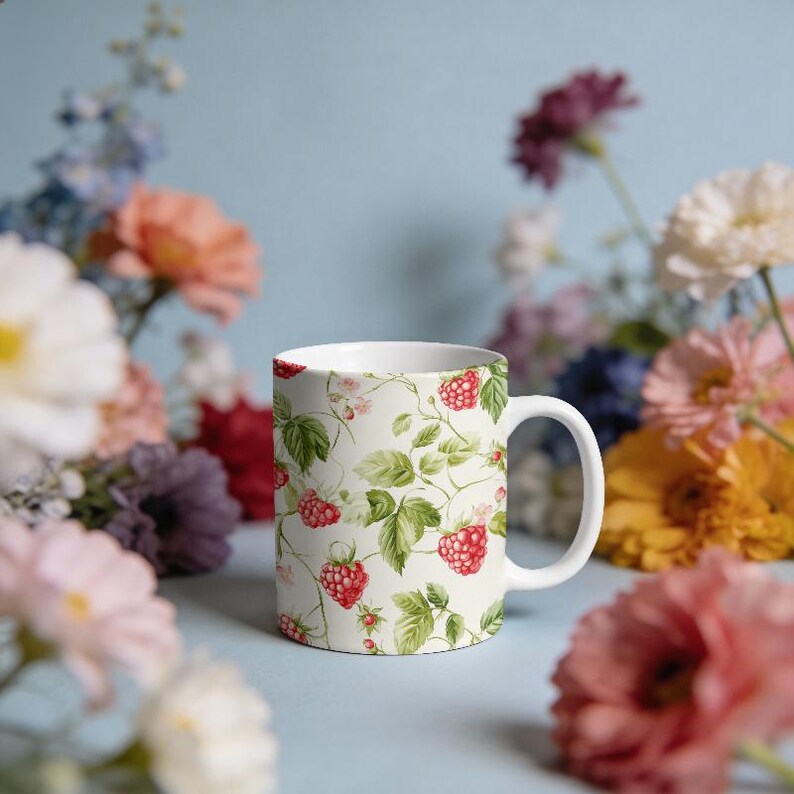 May include: White ceramic mug with a vibrant raspberry and leaf pattern. The design features red raspberries and green leaves on a cream background. The mug has a white handle and is surrounded by colorful flowers.