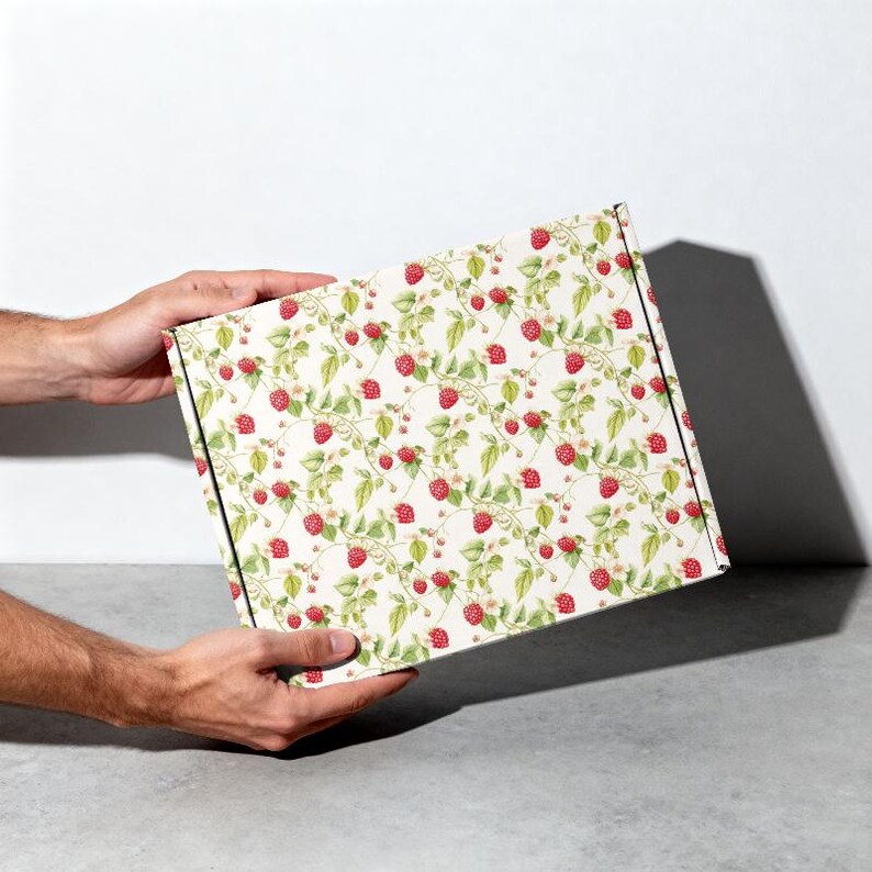 May include: A rectangular box with a repeating pattern of red raspberries, green leaves, and white flowers on a cream background. The box is held by two hands against a white and gray backdrop.