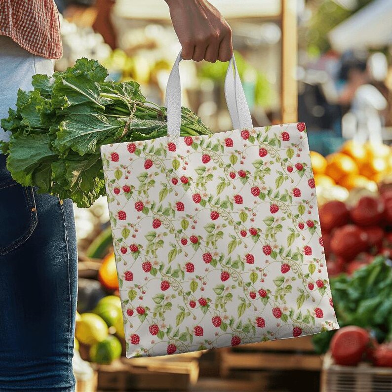 May include: A white tote bag with a repeating pattern of red raspberries, green leaves, and delicate white flowers. The bag has white handles and is being held near a farmer's market. The bag is filled with groceries.