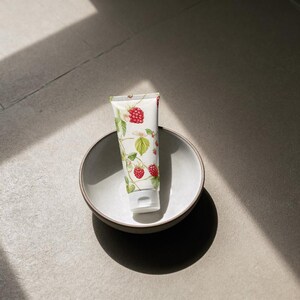 May include: A white tube of cream with a botanical raspberry design, resting in a gray ceramic bowl. The cream tube features red raspberries, green leaves, and white flowers. The bowl is round and has a simple, minimalist design.