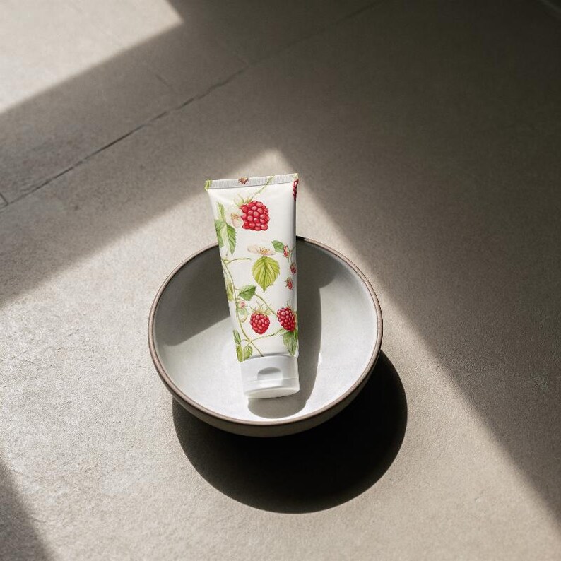May include: A white tube of cream with a botanical raspberry design, resting in a gray ceramic bowl. The cream tube features red raspberries, green leaves, and white flowers. The bowl is round and has a simple, minimalist design.
