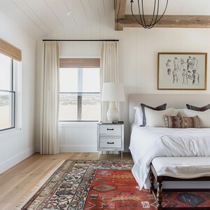Op de afbeelding: Een slaapkamer met een wit bed, nachtkastje en gordijnen. Een rood tapijt met patroon bedekt de vloer. Een ingelijst kunstwerk hangt boven het bed. De kamer heeft een neutraal kleurenpalet met natuurlijk licht.