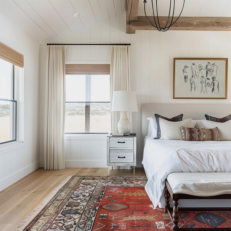 Op de afbeelding: Een slaapkamer met een wit bed, nachtkastje en gordijnen. Een rood tapijt met patroon bedekt de vloer. Een ingelijst kunstwerk hangt boven het bed. De kamer heeft een neutraal kleurenpalet met natuurlijk licht.