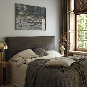 May include: A bedroom with a dark brown headboard, a bed with white linens and a gray throw blanket, a small wooden nightstand with a gold lamp, and a framed watercolor painting of a river scene above the headboard.
