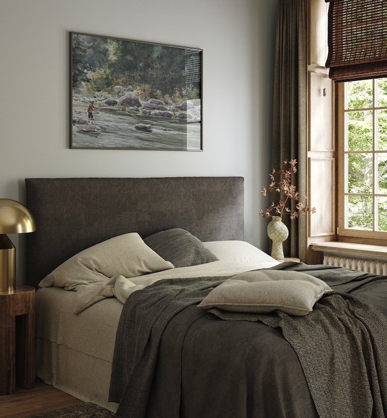 May include: A bedroom with a dark brown headboard, a bed with white linens and a gray throw blanket, a small wooden nightstand with a gold lamp, and a framed watercolor painting of a river scene above the headboard.