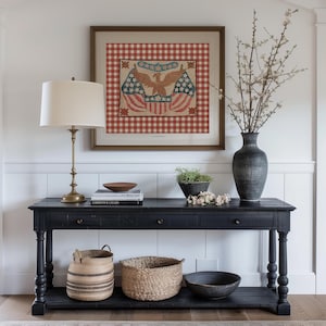 Puede incluir: Una consola negra con una obra de arte enmarcada con un &aacute;guila y banderas. Una l&aacute;mpara de lat&oacute;n, libros y cuencos decorativos est&aacute;n sobre la mesa. Cestas tejidas y un jarr&oacute;n oscuro con ramas completan la decoraci&oacute;n. La obra tiene un fondo a cuadros rojos y blancos.