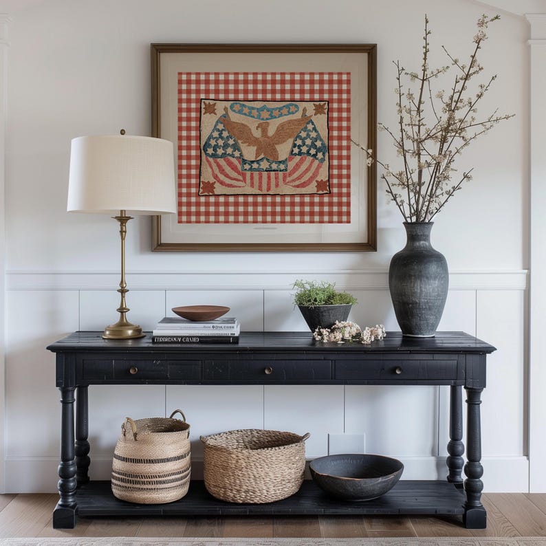 Puede incluir: Una consola negra con una obra de arte enmarcada con un &aacute;guila y banderas. Una l&aacute;mpara de lat&oacute;n, libros y cuencos decorativos est&aacute;n sobre la mesa. Cestas tejidas y un jarr&oacute;n oscuro con ramas completan la decoraci&oacute;n. La obra tiene un fondo a cuadros rojos y blancos.