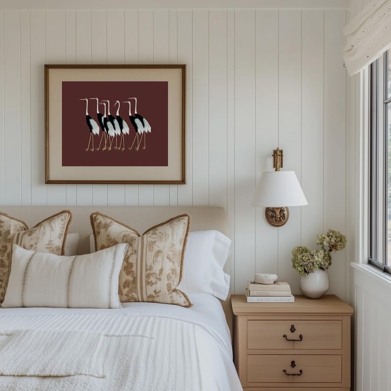 May include: A white bed with a white and brown patterned throw blanket and a white and brown patterned pillow. There is a framed print of black and white birds on a maroon background above the bed. A nightstand with three drawers is on the right side of the bed.