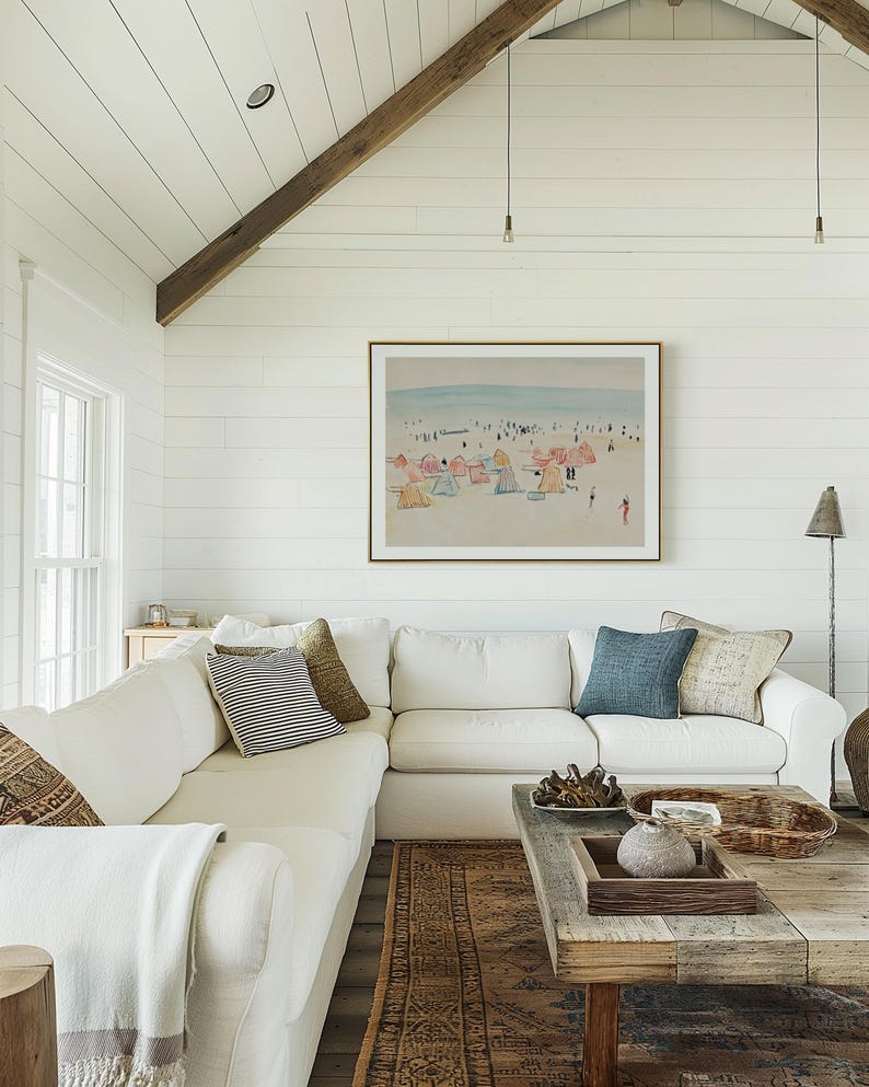 May include: A white sectional sofa with throw pillows and a white throw blanket sits in front of a white shiplap wall with a framed watercolor painting of a beach scene. A wooden coffee table with a woven basket and a decorative stone sits in front of the sofa. A brown patterned rug is on the floor.