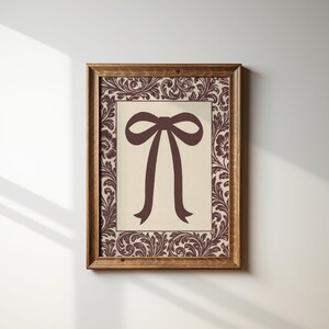 May include: A framed print featuring a brown bow illustration on a cream background. The print is surrounded by a decorative border with floral and scroll patterns in brown. The frame is made of wood.