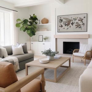 May include: A modern living room with a neutral color palette. A large framed artwork of birds hangs above a fireplace. A gray sofa, wooden coffee table, and woven chair are in the room. A large potted plant adds a touch of greenery.