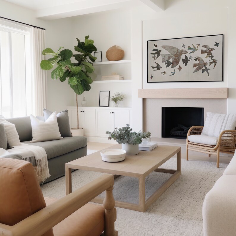 May include: A modern living room with a neutral color palette. A large framed artwork of birds hangs above a fireplace. A gray sofa, wooden coffee table, and woven chair are in the room. A large potted plant adds a touch of greenery.
