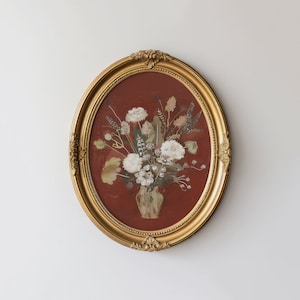 Puede incluir: Un marco ovalado dorado con fondo rojo y un arreglo de flores prensadas en un jarrón. Las flores son blancas y beige y el jarrón es marrón.