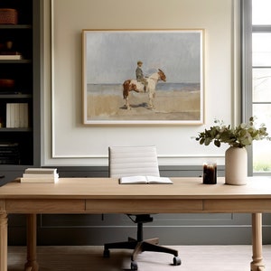 May include: A light wood desk with a white office chair and a framed painting of a person riding a horse on a beach. There is a vase with green leaves and a candle on the desk.
