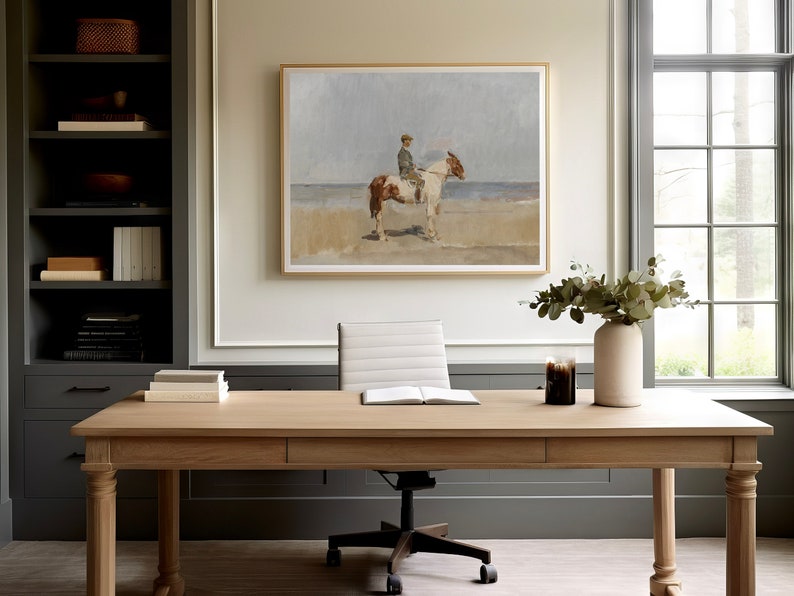 May include: A light wood desk with a white office chair and a framed painting of a person riding a horse on a beach. There is a vase with green leaves and a candle on the desk.