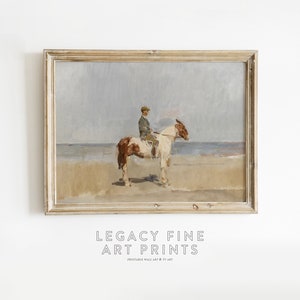 May include: A framed painting of a person riding a brown and white horse on a beach. The sky is a light blue and the water is a light gray. The painting is in a light wood frame.