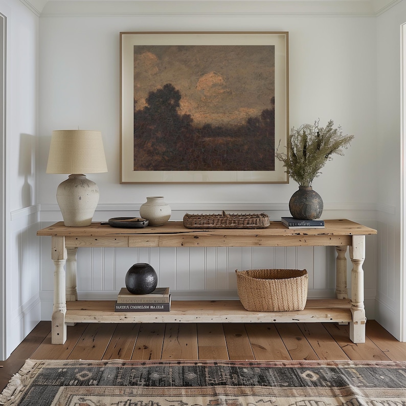 May include: A rustic wooden console table with a whitewashed finish features a framed landscape painting, a ceramic lamp, a woven basket, and a collection of books. The table has two shelves and is styled with decorative objects.